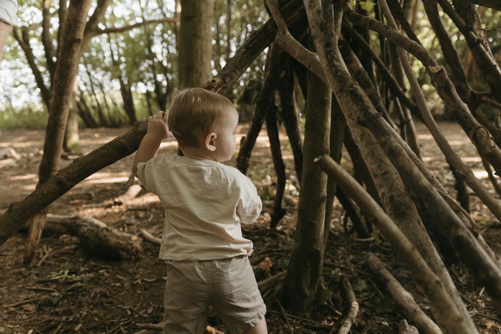A small child wearing beige clothing explores a lean-to structure made of sticks and branches in a wooded area.