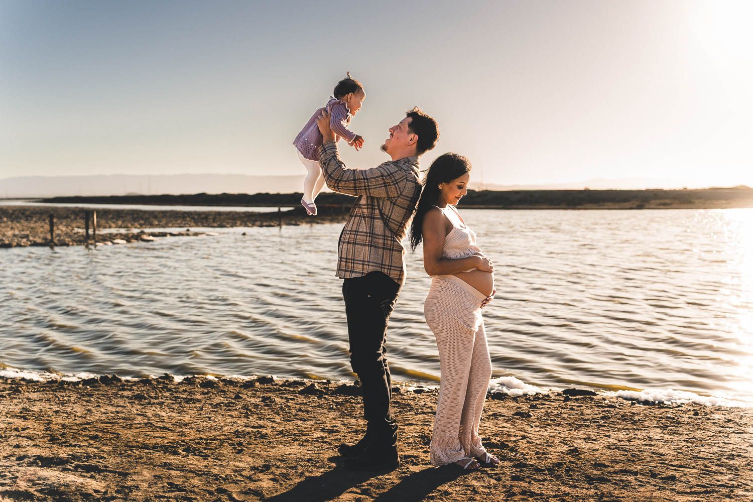 A family shares a tender moment on the beach at sunset, standing by the water's edge.