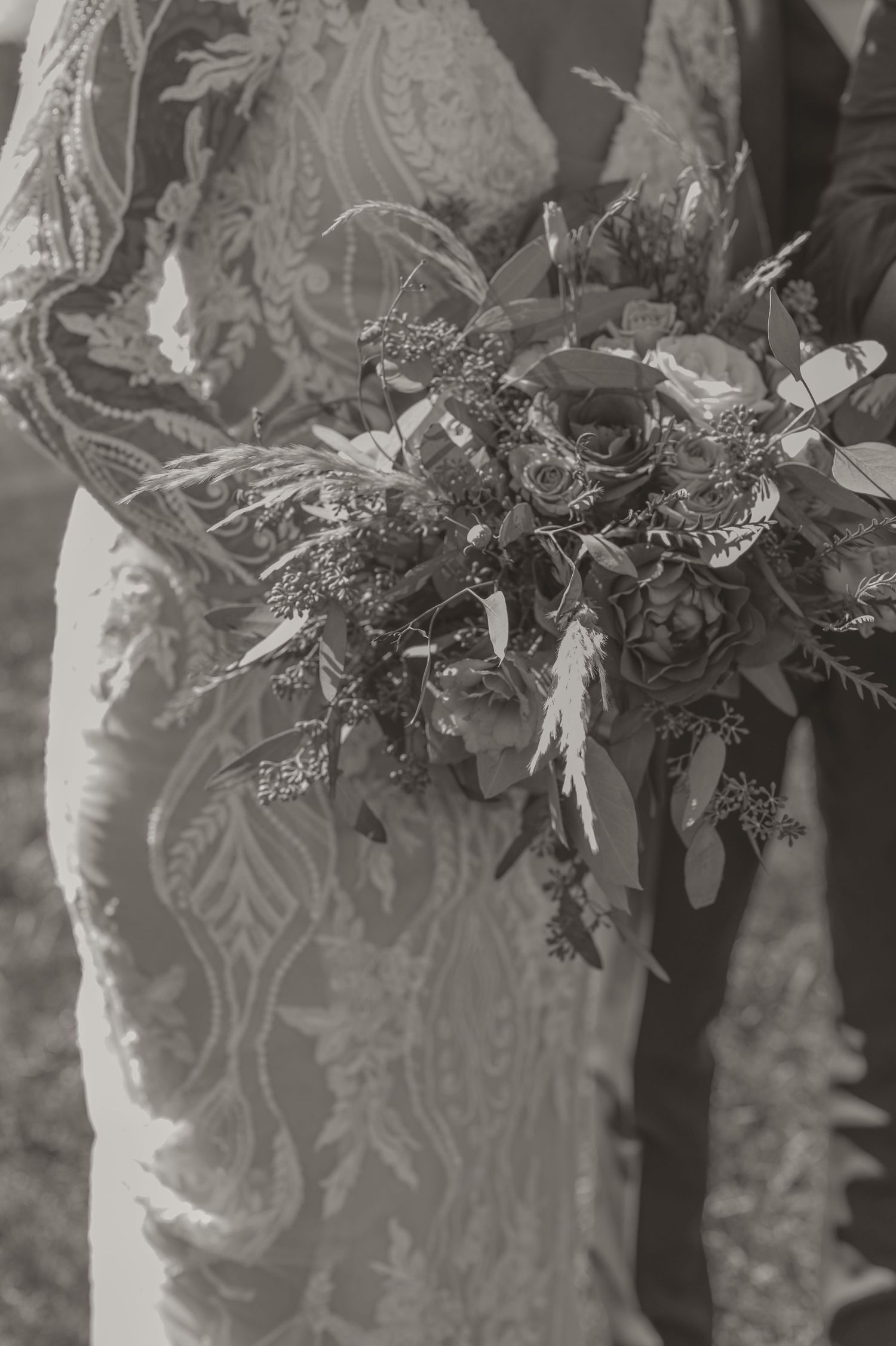 A detailed black and white photograph by Samantha Campbell Imagery showing an ornate lace wedding dress and rustic wildflower bouquet.