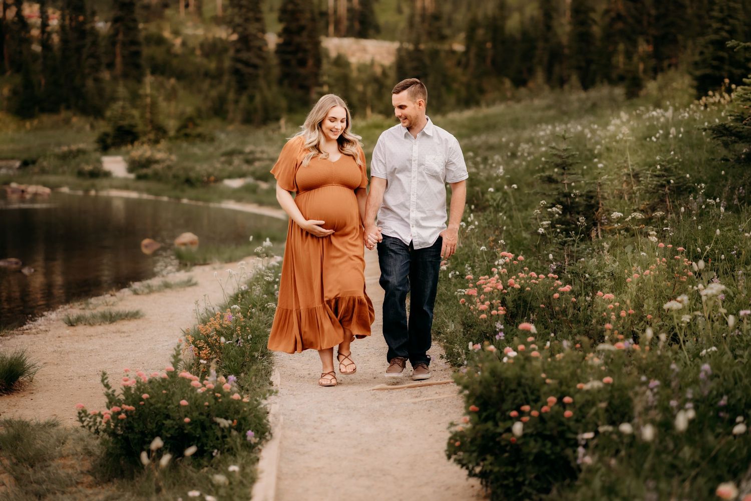 Husband and wife walk hand in hand aside Tipsoo Lake surrounded by wildflowers for their maternity photo session in the mountains.