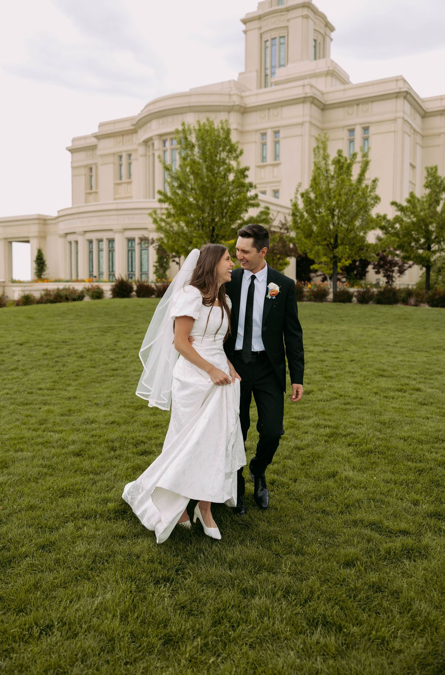 A joyful wedding couple exits a building with white flowers and elegant doors.