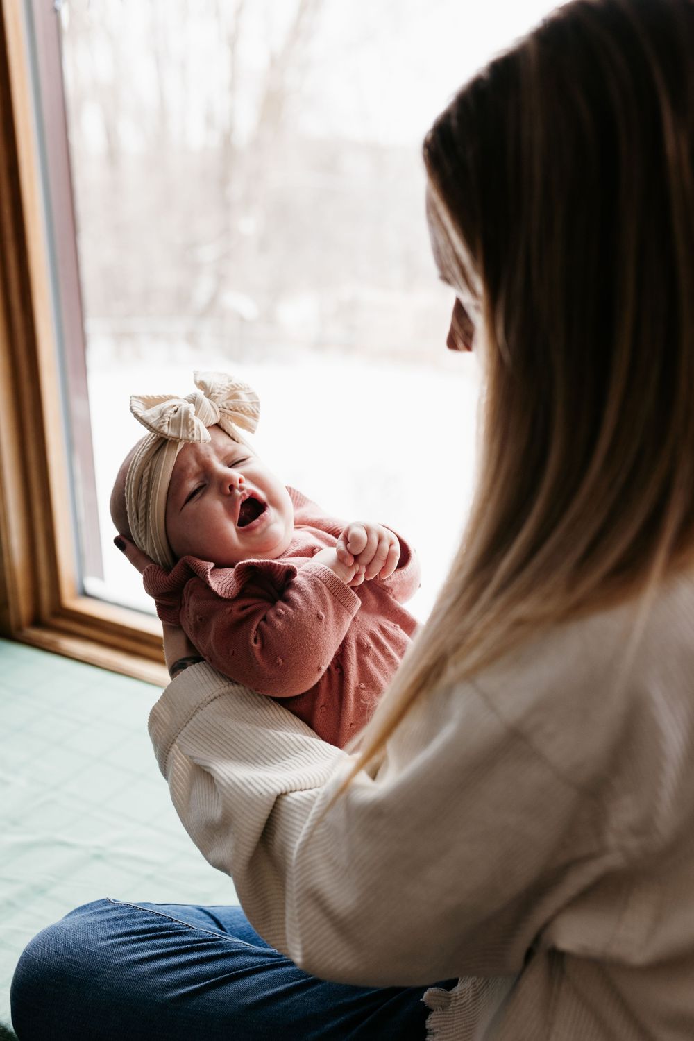 A baby in pink clothing and headband smiles with joy while being held near a window.