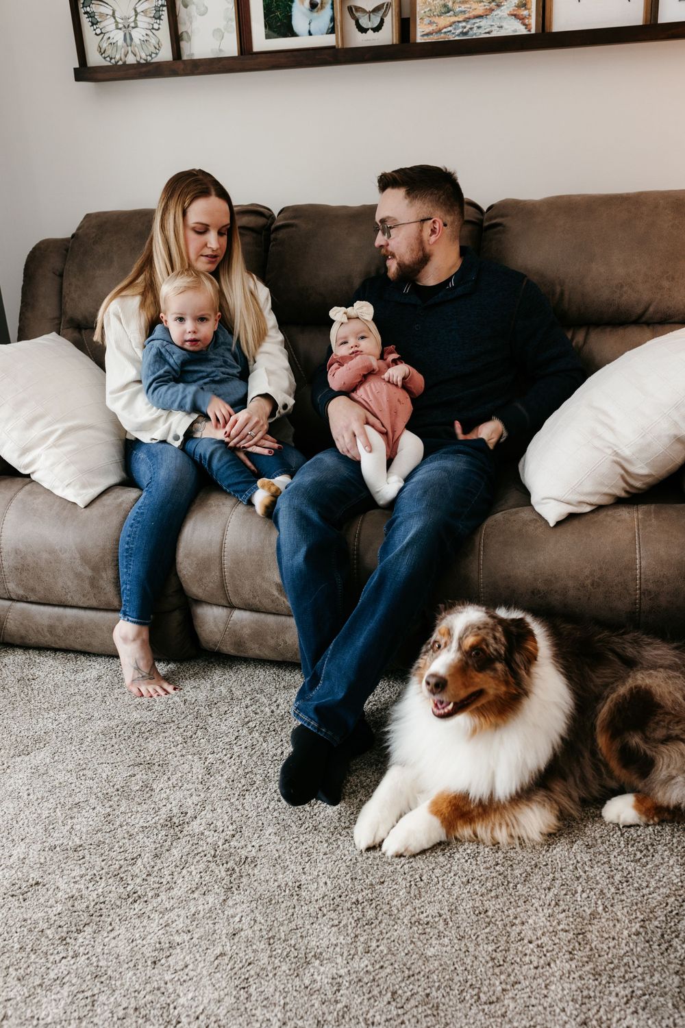 Young couple sitting on brown couch with two small children and Australian Shepherd dog resting on carpet below.