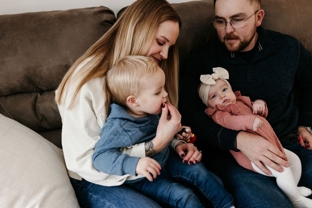 Family sits together on brown couch with toddler in denim and newborn baby in pink during intimate home session.