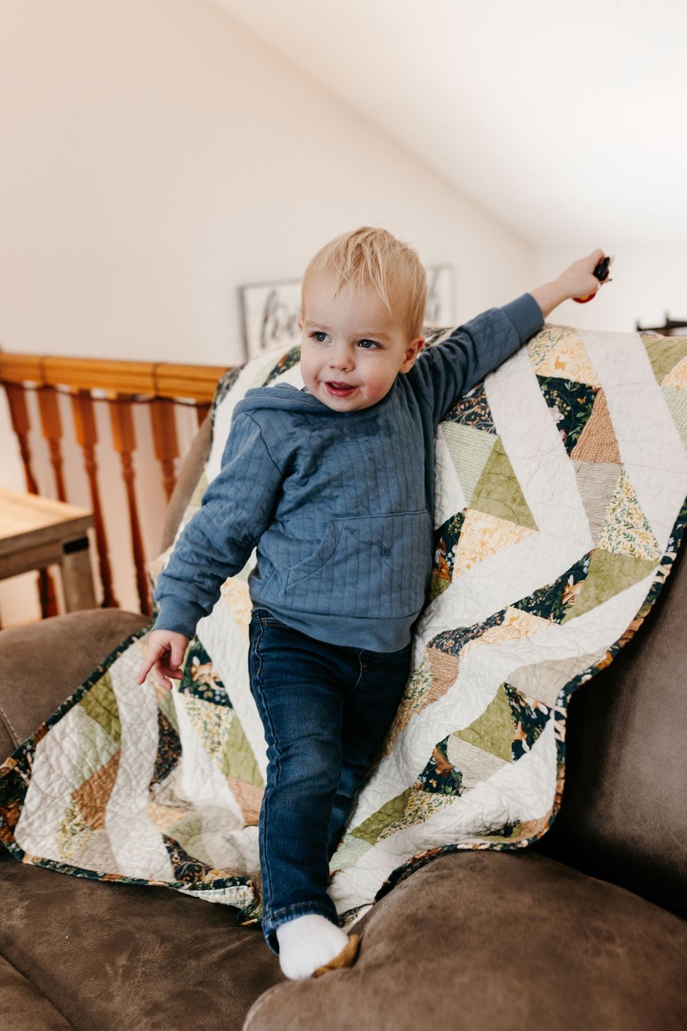 A joyful toddler in a blue sweater and jeans stands on a couch next to a patterned geometric quilt.