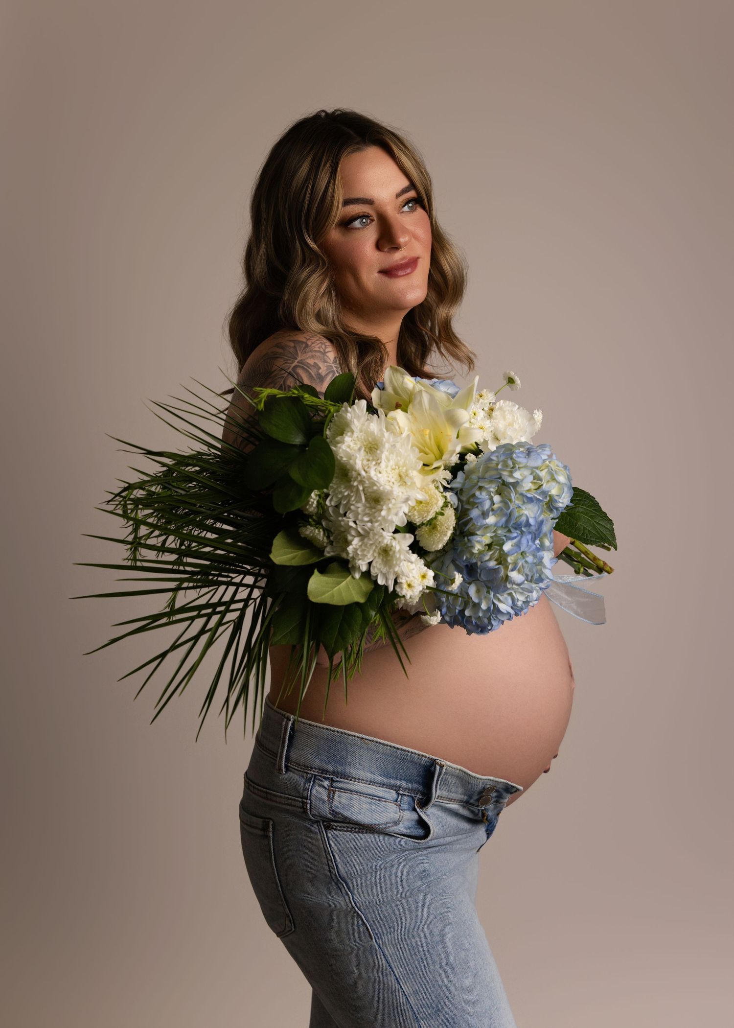 Pregnant woman in jeans with bare belly holding a bouquet of white and blue hydrangeas against beige background.