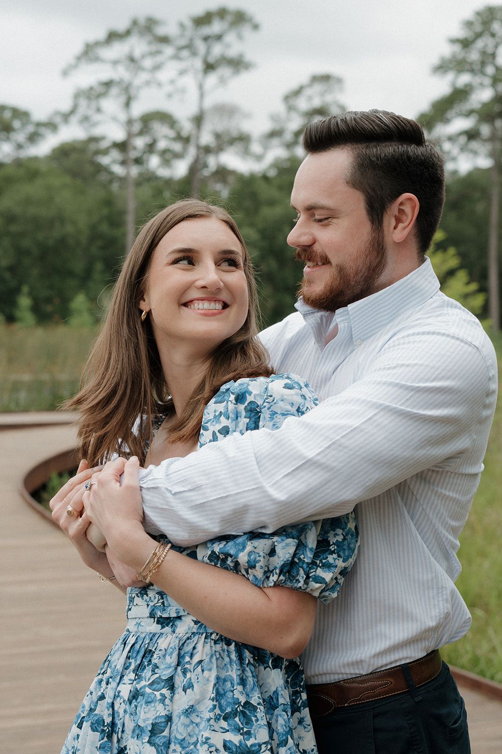 Couple embracing outdoors on wooden boardwalk with blue floral dress and white shirt against pine tree background.