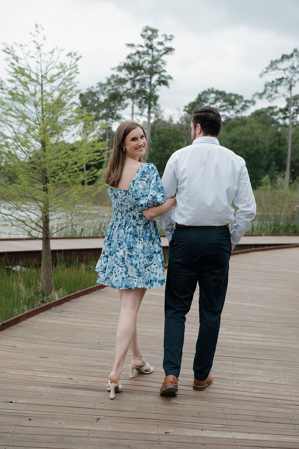 A couple walks along a wooden boardwalk surrounded by trees and greenery, with her wearing a blue floral dress.