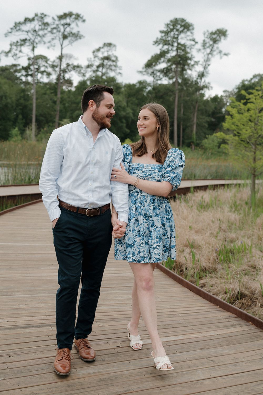 A couple walks hand in hand on a wooden boardwalk surrounded by tall pine trees and marsh grass on a cloudy day.