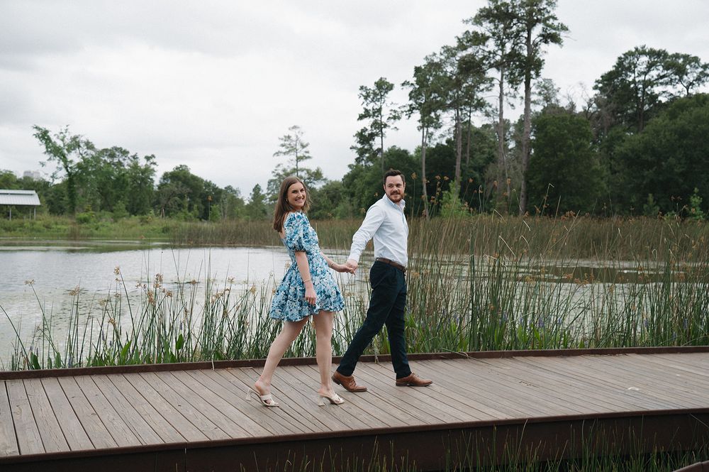 Couple walking hand in hand along a wooden boardwalk beside a serene pond surrounded by tall pine trees.
