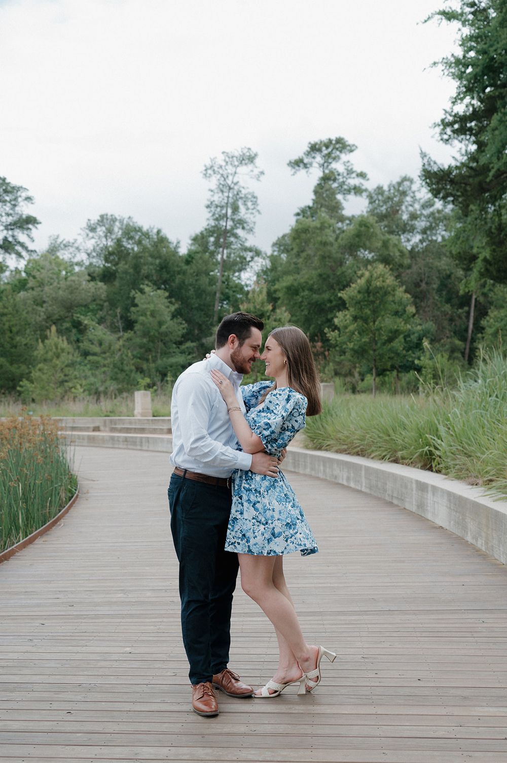 A romantic couple embraces on a wooden boardwalk surrounded by lush greenery and natural landscaping.