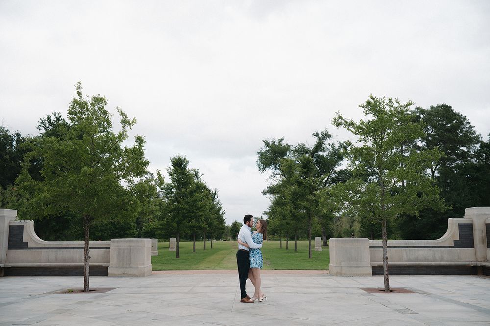 A couple embraces on a tree-lined path between concrete barriers at a park on an overcast day.