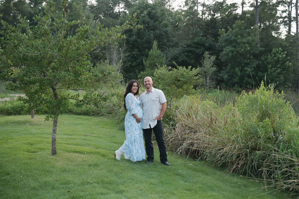 Couple in coordinated light blue and white outfits embrace on a lawn near ornamental grasses at sunset.