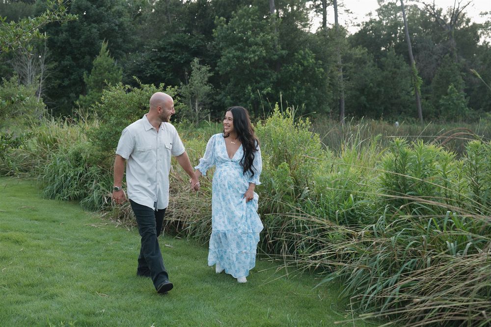Two people take a romantic stroll along a garden pathway next to tall grass and greenery on a summer evening.
