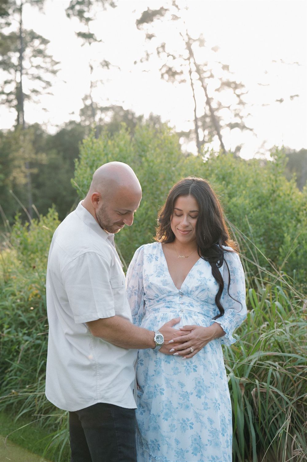 Couple in outdoor setting with tall grass and trees as woman in blue floral dress cradles her baby bump.