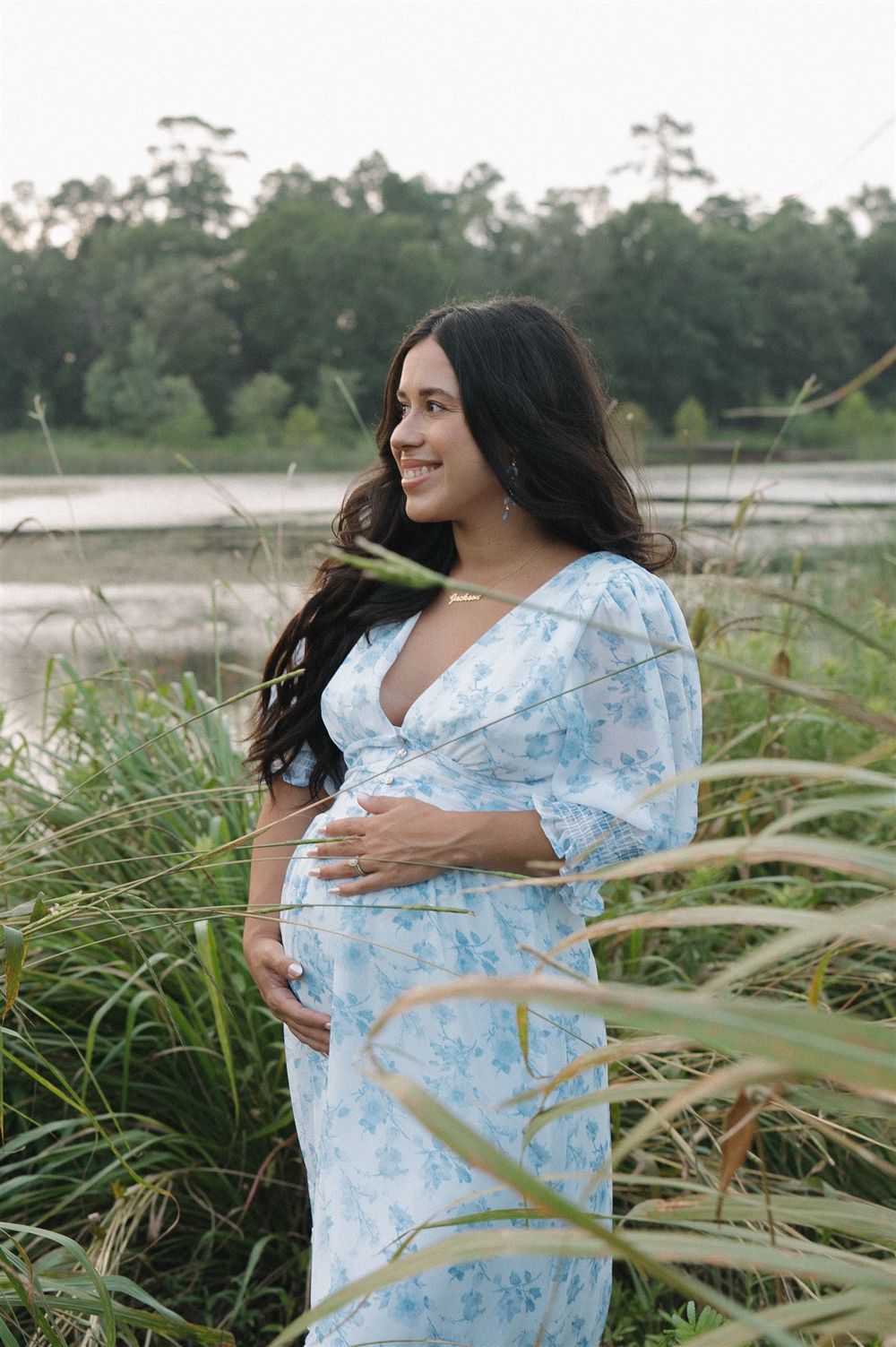 Expecting mother in a light blue floral dress poses in tall grass by a lake during a maternity photoshoot.