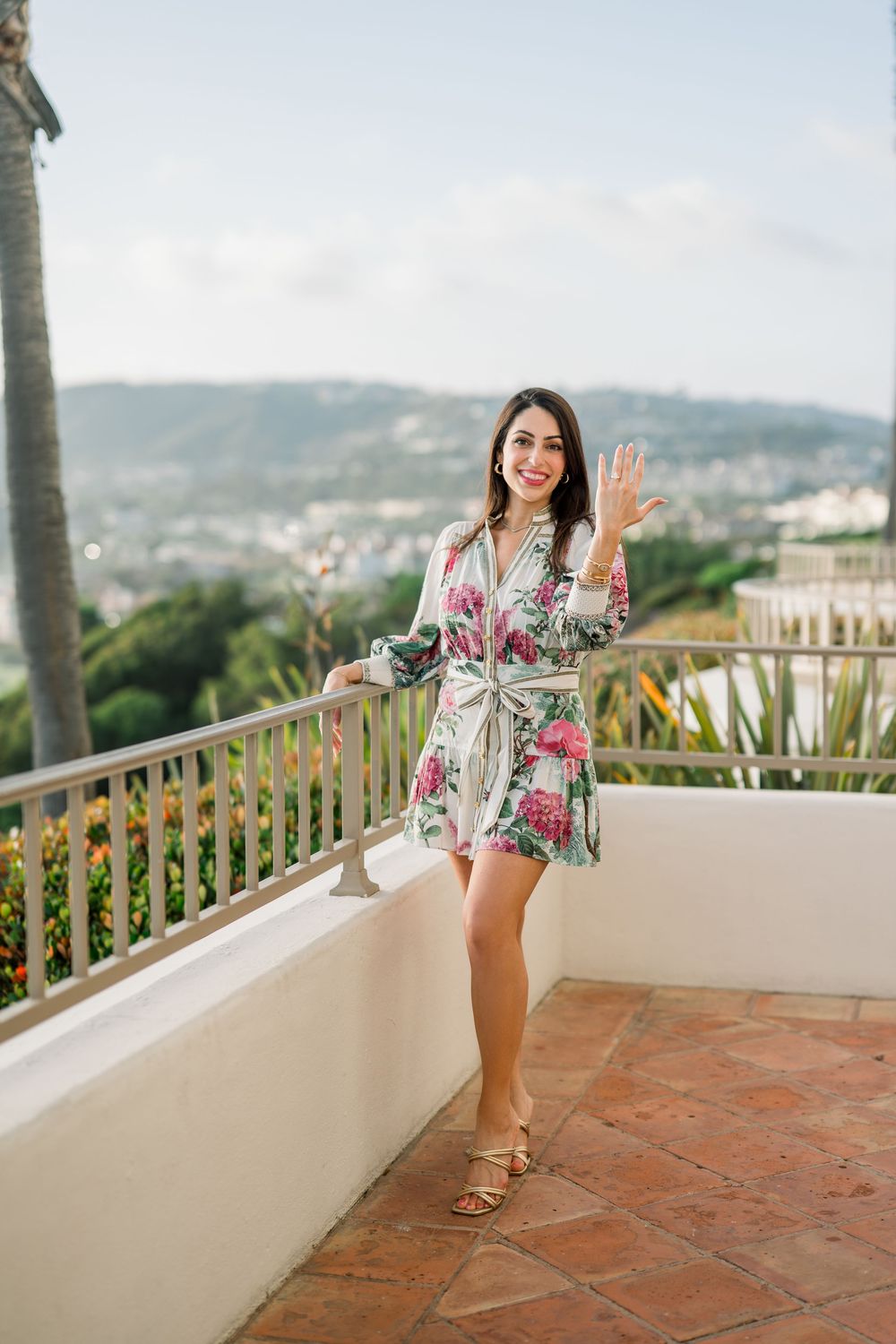 A person in a floral robe stands on a tropical balcony with mountain views, waving and smiling on terracotta tiles.
