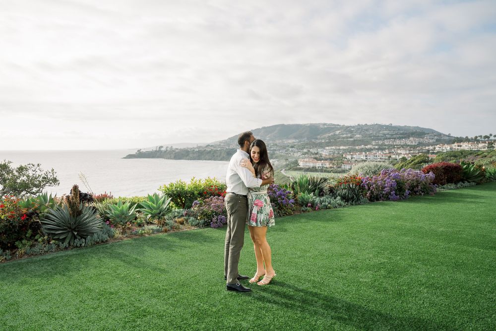 Couple embraces on lush green lawn overlooking coastal bay with colorful flowers and ocean view in background.
