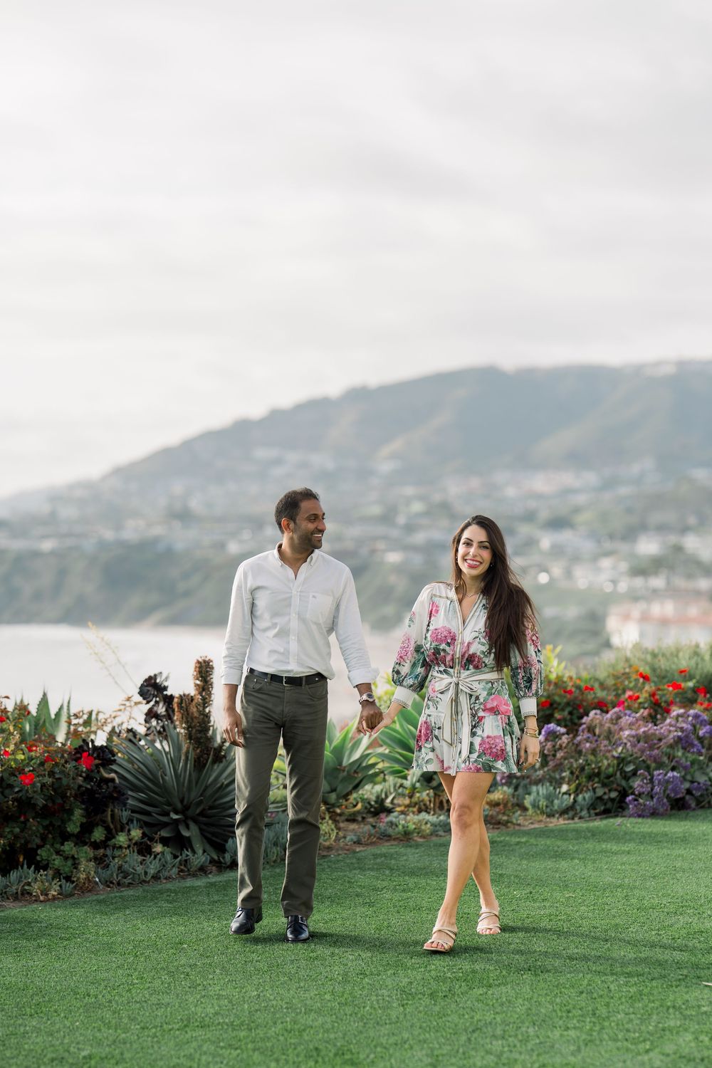 A couple strolling together on a landscaped lawn with flowering gardens and coastal mountains in the background.
