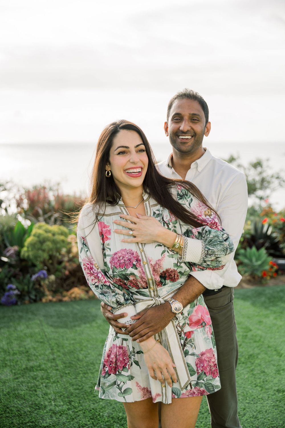 A couple embraces and laughs together outdoors in a garden, with the woman wearing a floral print dress.