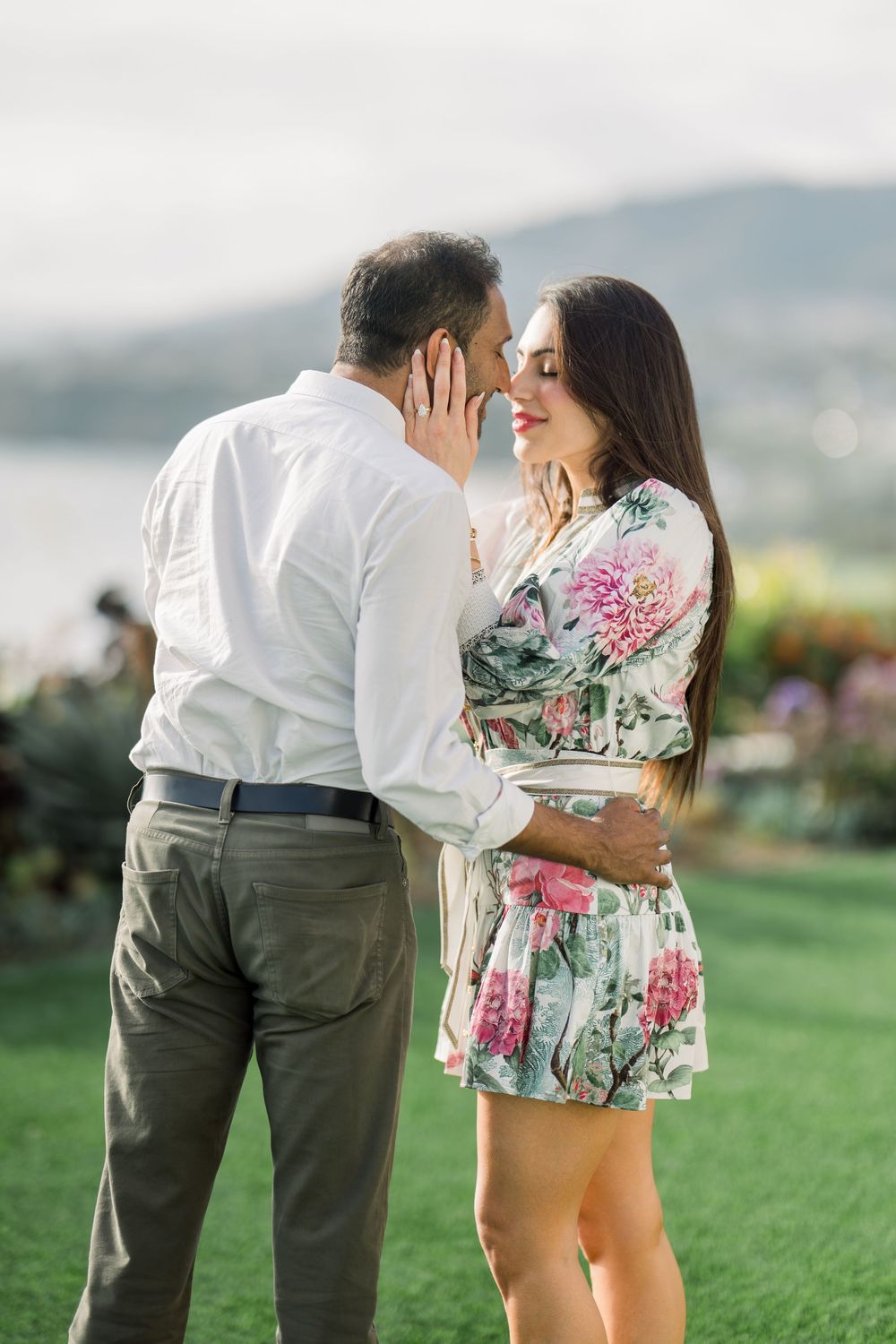 A romantic couple embraces on a grassy hilltop overlooking mountains, with the woman wearing a floral dress and man in white shirt.