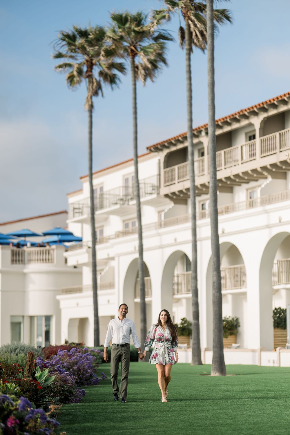 A couple walks along a green lawn path lined with palm trees and Spanish colonial architecture at a luxury resort.