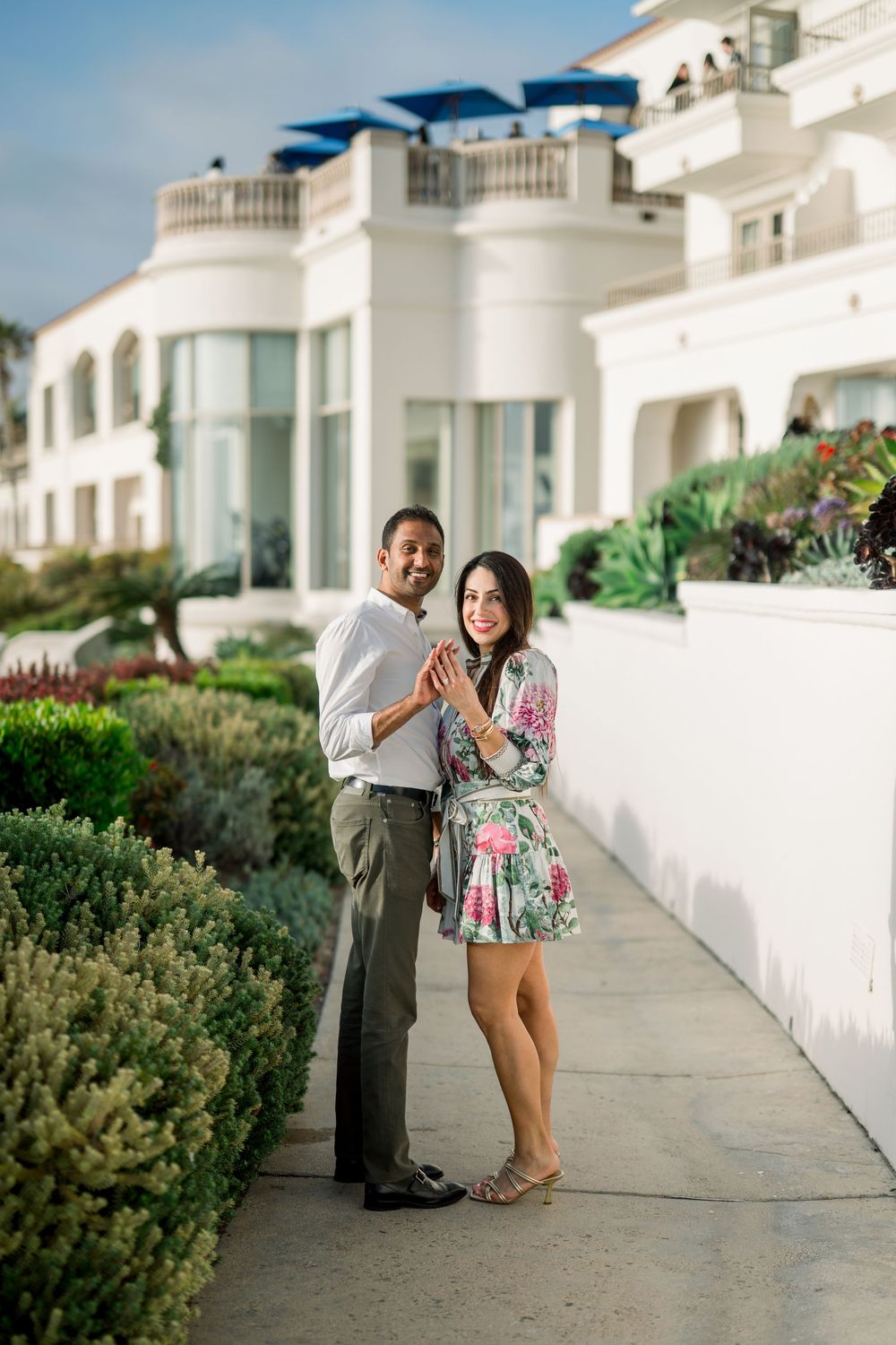 A couple poses together on a white walkway outside a Mediterranean-style building with blue awnings and lush gardens.