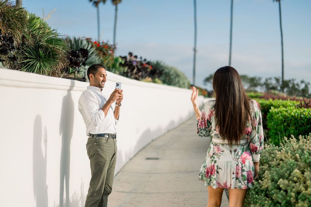 A couple stands along a scenic white wall walkway with palm trees and flowering plants on a sunny day.