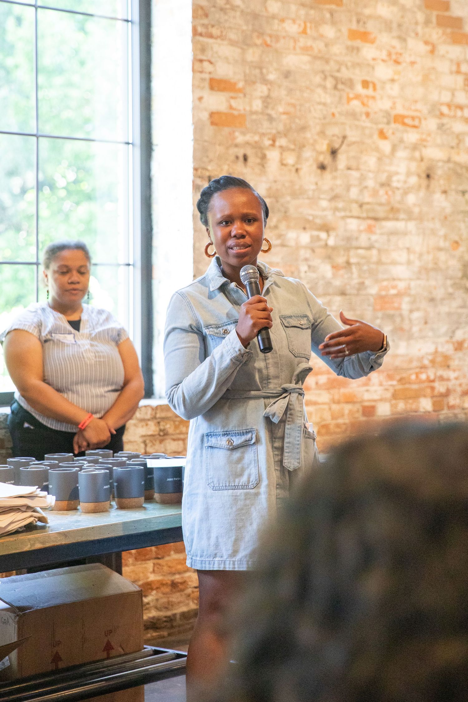 Someone speaks into a microphone while wearing denim at a presentation with a brick wall backdrop and coffee service behind.