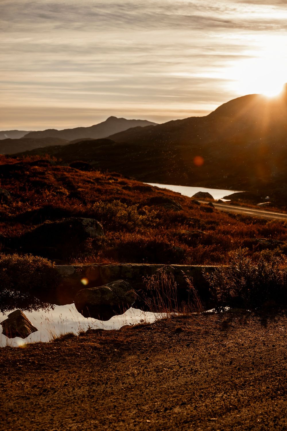 Dramatic sunset over mountain landscape with reflective water and rugged terrain in warm sepia tones.