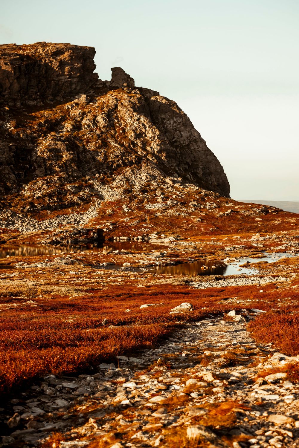 Dramatic rocky cliff face rises above rusty orange moorland with scattered stones and a winding path at sunset.