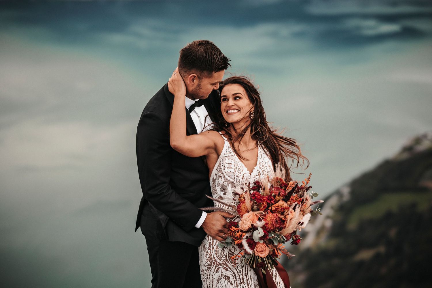 A couple in formal attire embrace on a cliffside while the bride holds a vibrant autumn-colored bouquet.