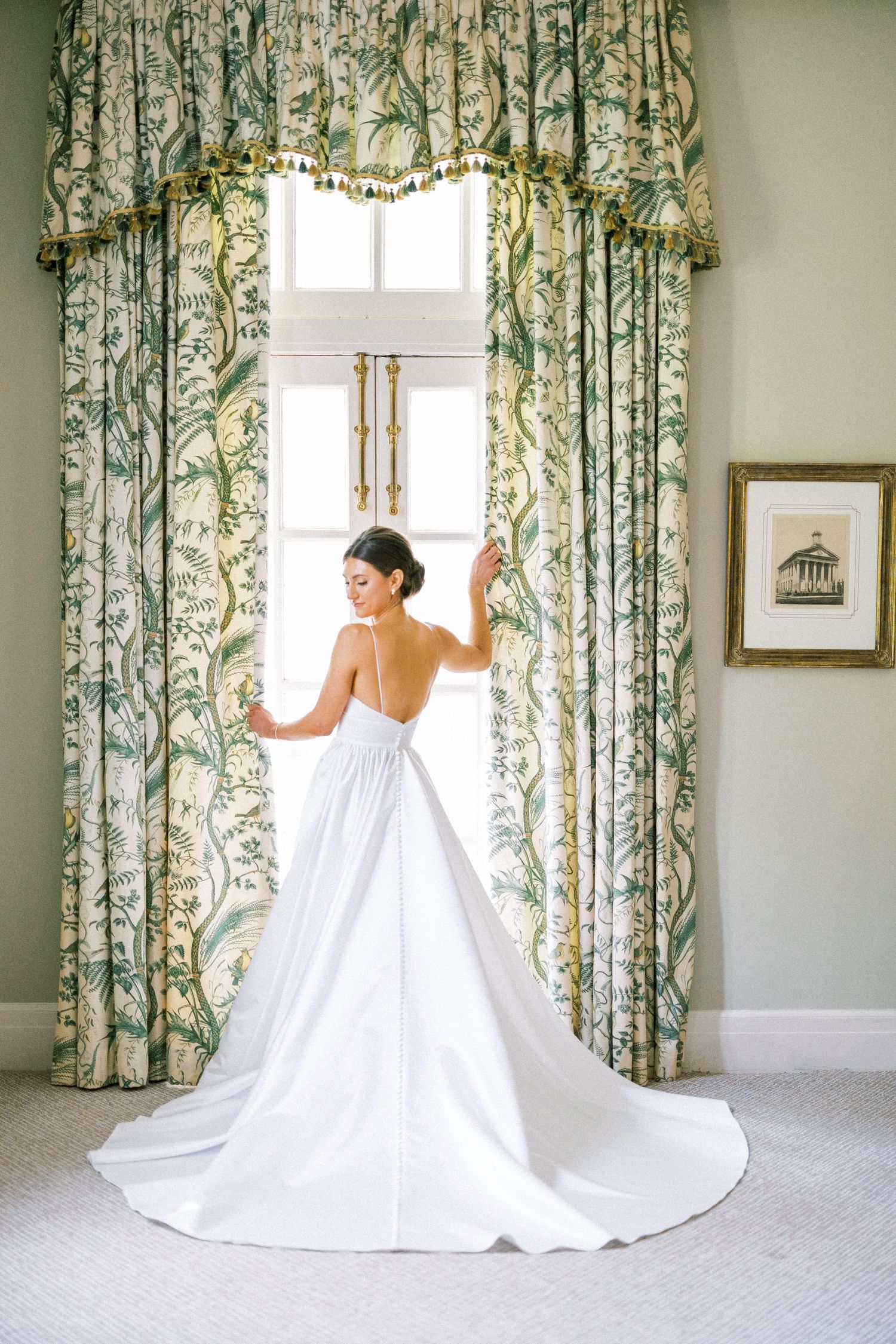 Elegant wedding dress with long train photographed against floral curtains in a classic interior.