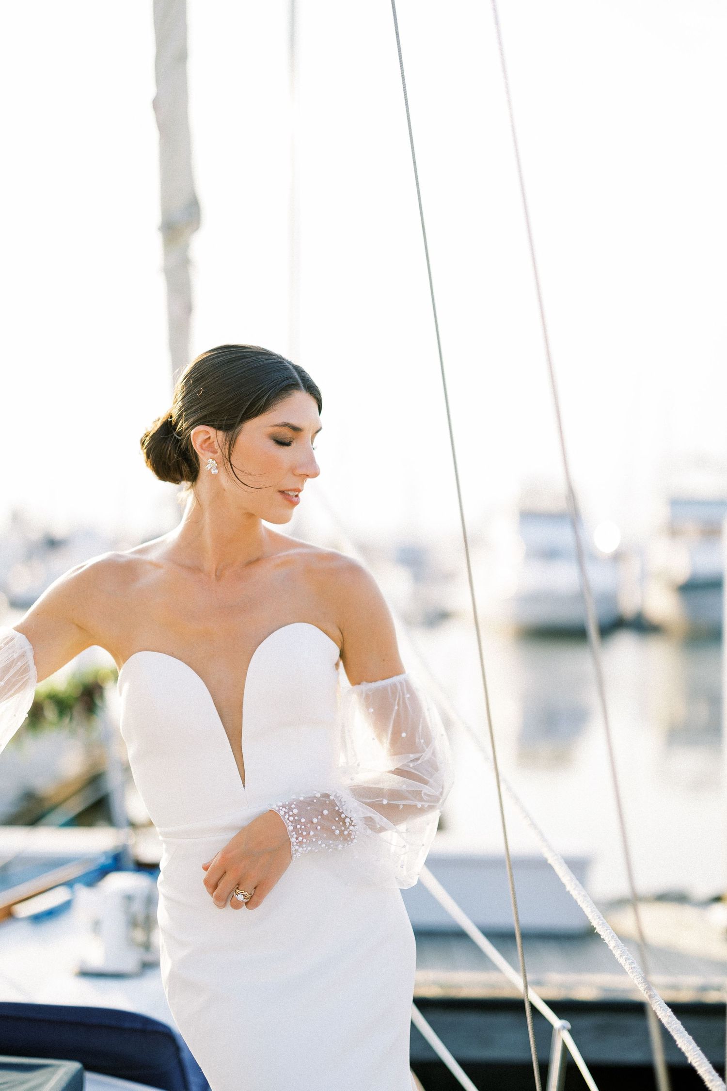 An elegant bride in a modern white wedding dress with sheer sleeves stands on a yacht in a marina during golden hour.