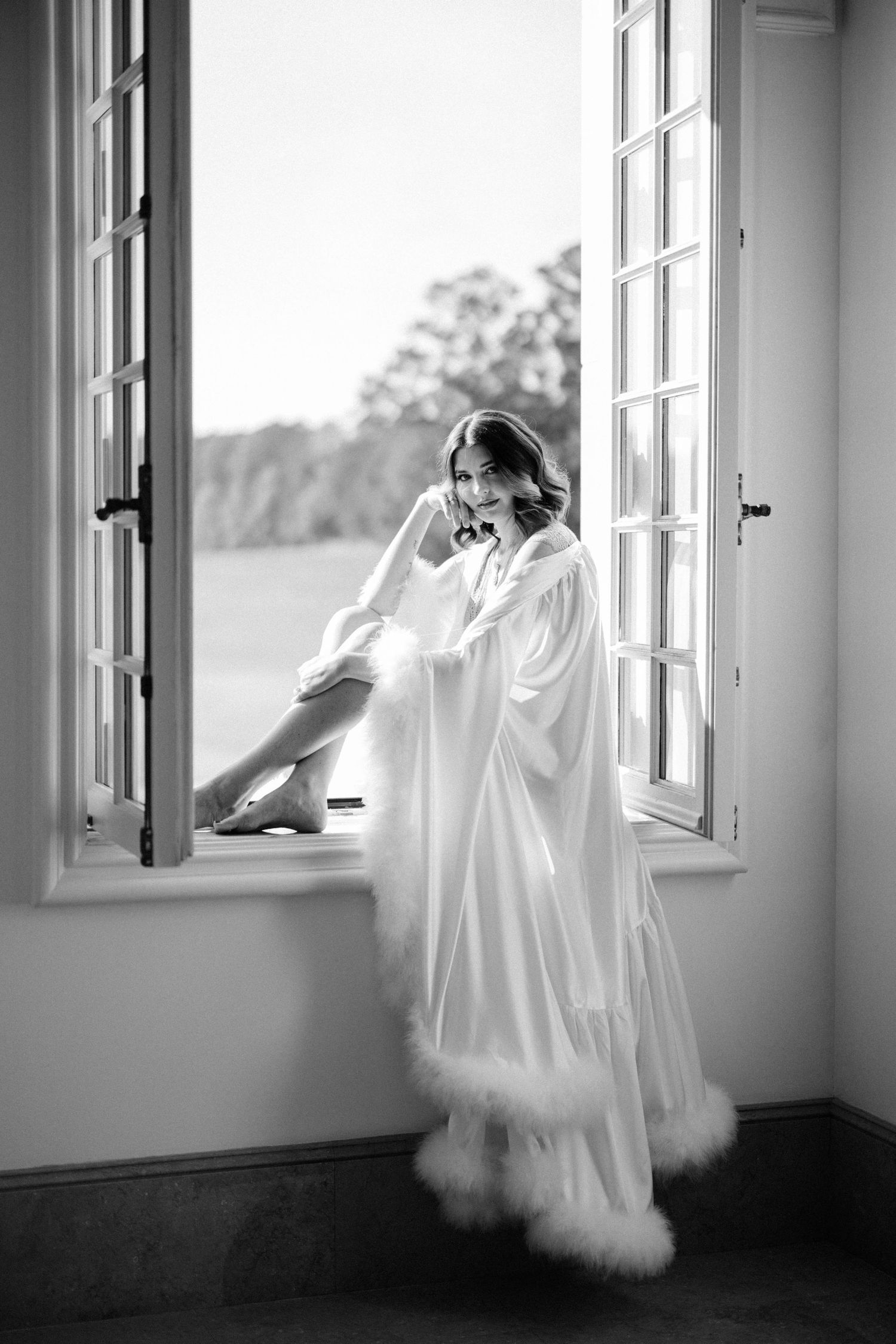 Elegant bridal portrait in white robe by window in natural light.