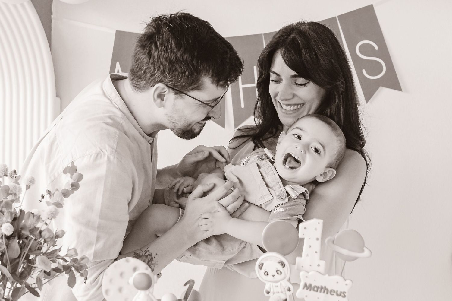 Playful black and white family moment showing parents tickling laughing baby while decorated banner hangs in background.