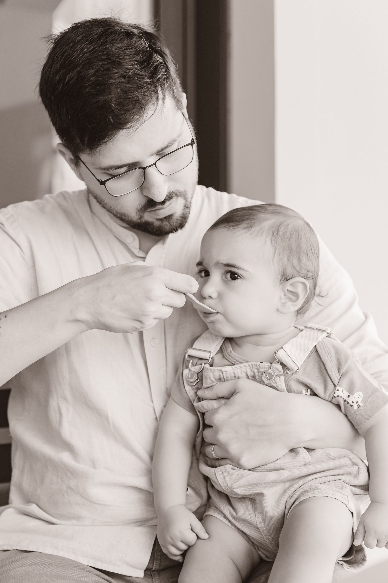 A person in glasses wearing a white shirt gently feeds a baby in a light-colored romper in this black and white photo.