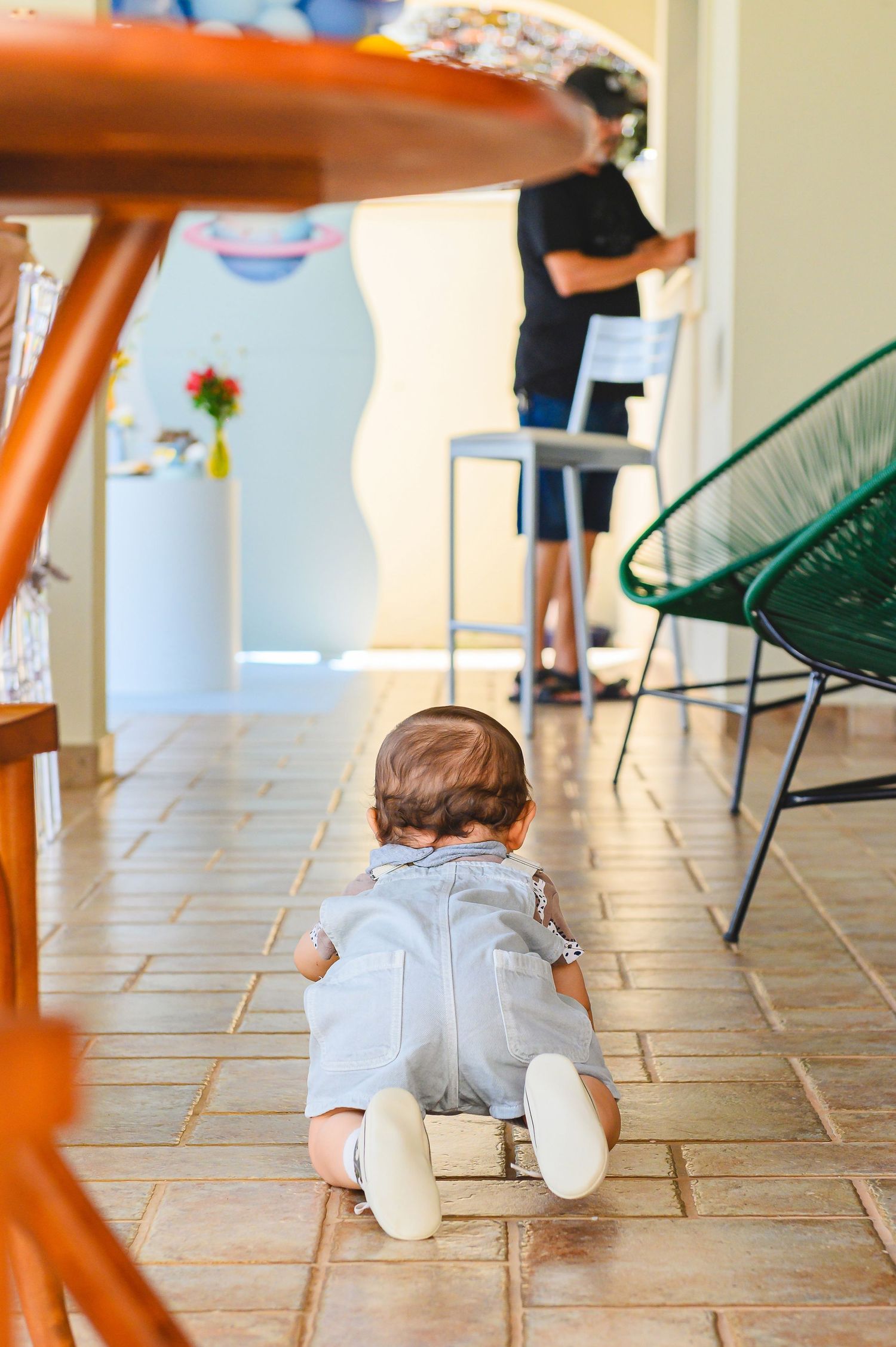 Small child in gray onesie crawling on tile floor while someone stands in background of sunny kitchen space.