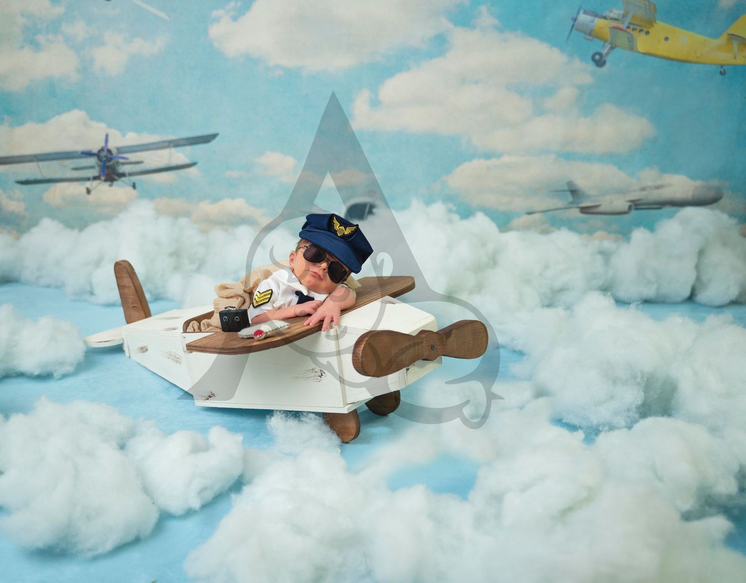 Whimsical newborn photography setup featuring a vintage-style airplane prop among painted clouds and toy planes.