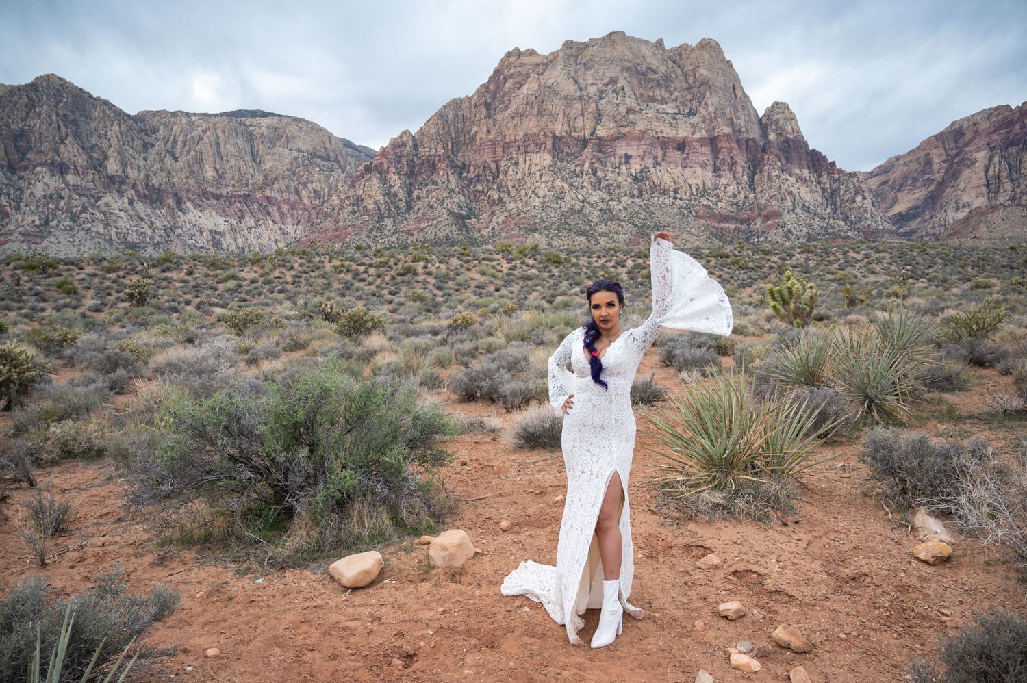 Bride strikes a fierce pose in a lace gown and boots, set against a dramatic desert mountain backdrop under a cloudy sky.