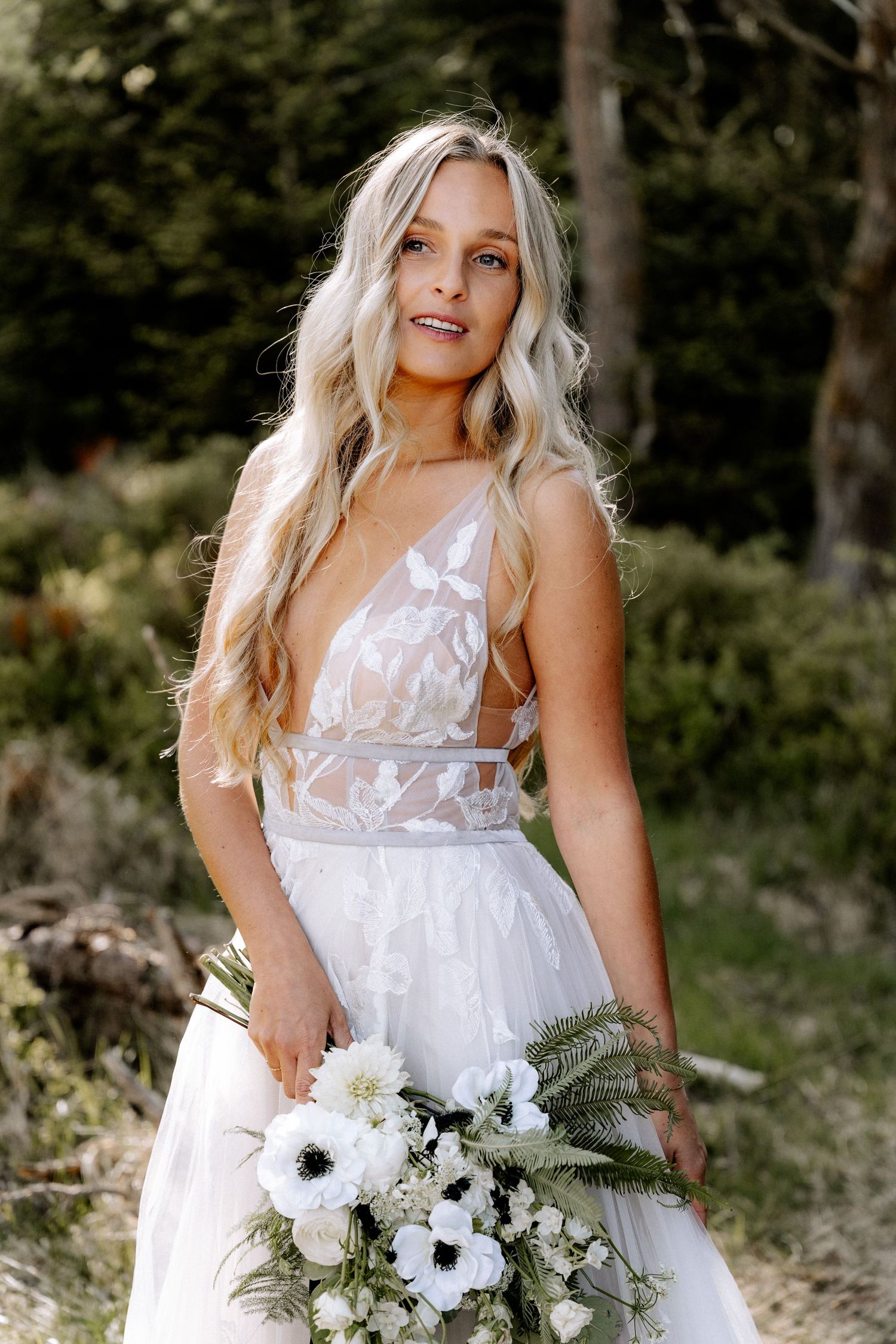 A bride in a delicate white lace wedding gown holds a bouquet of white anemone flowers and greenery in an outdoor setting.