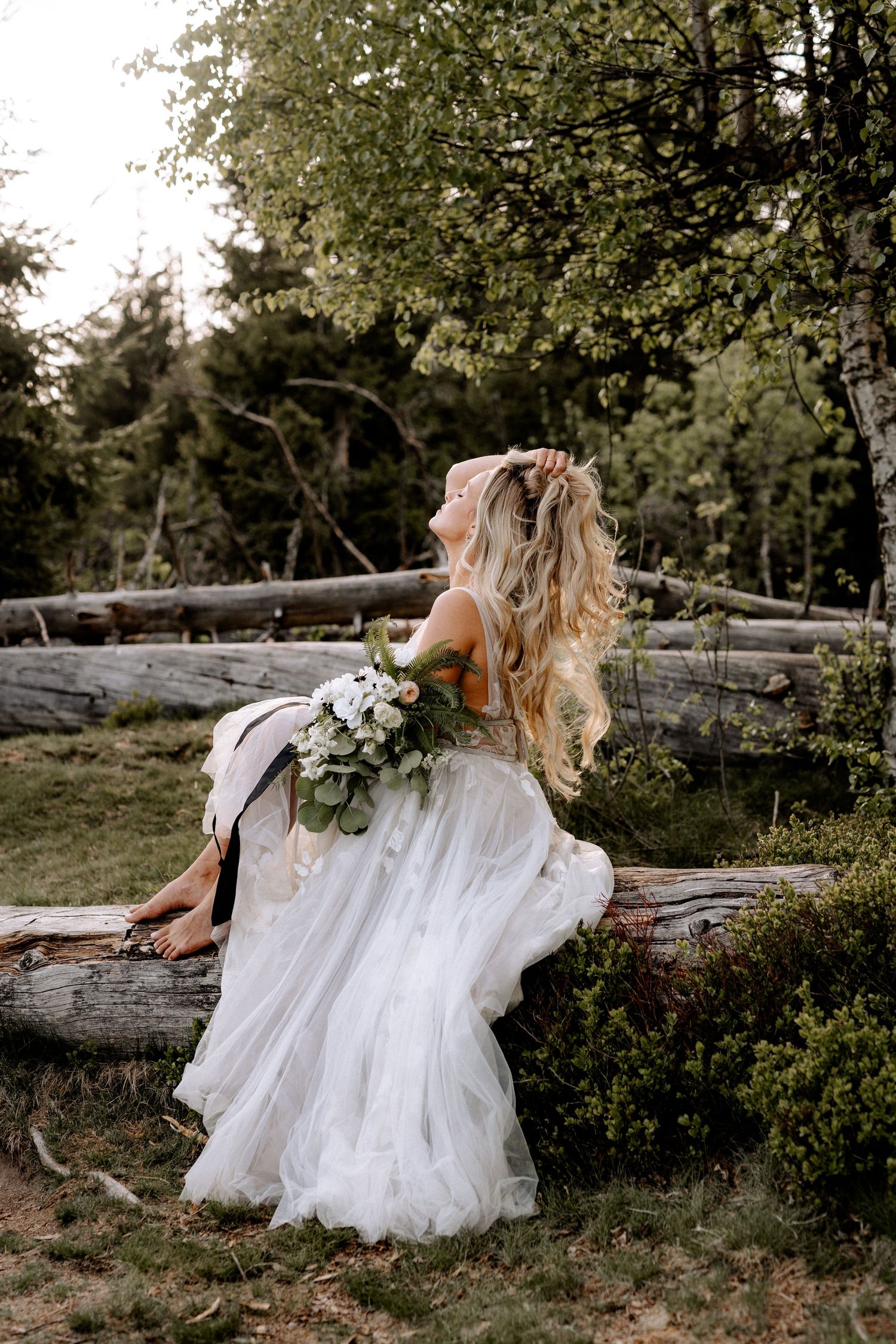 Dreamy bride in flowing white gown sits on rustic log holding bouquet with blonde hair flowing in outdoor forest setting.