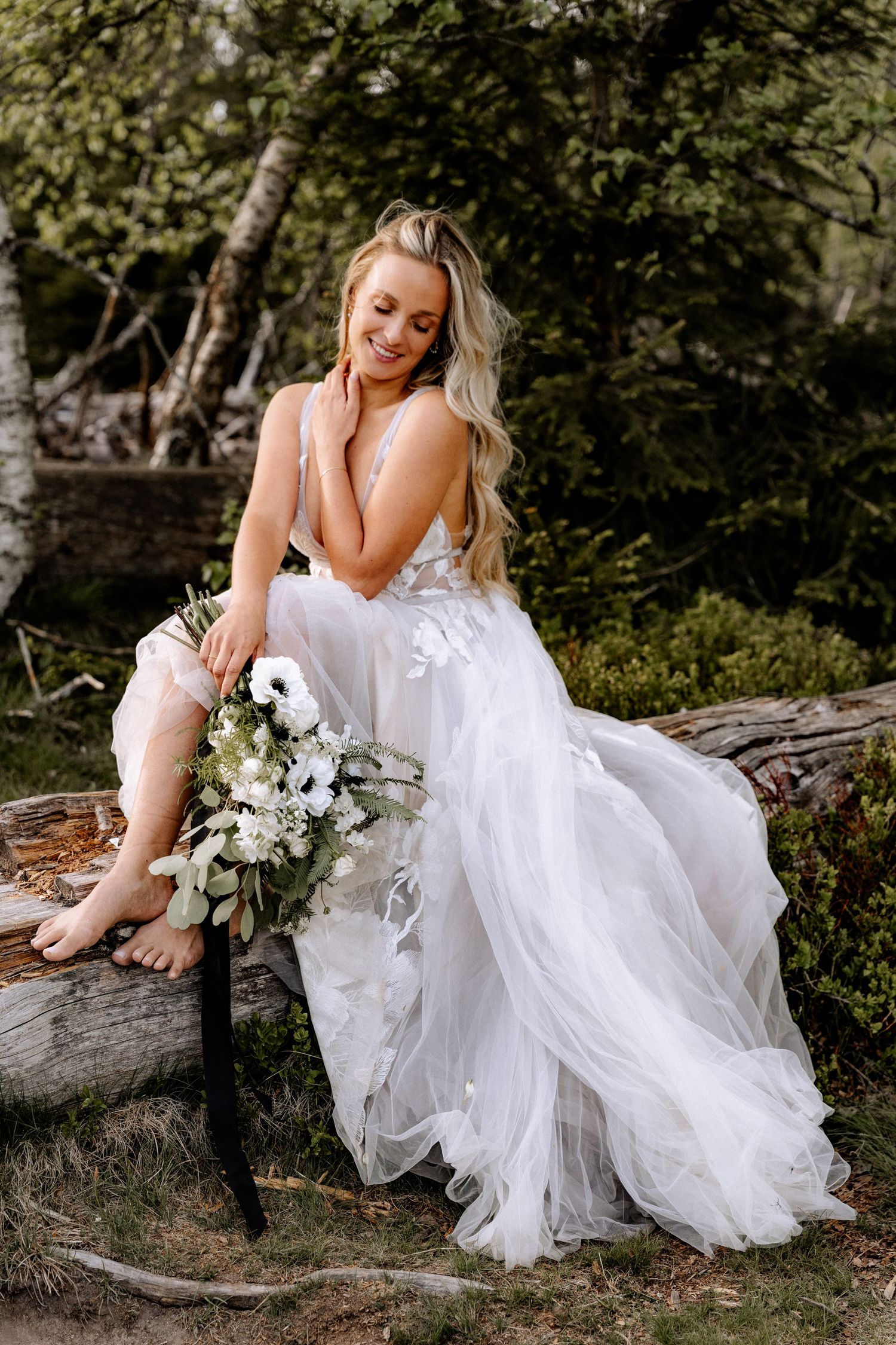 Bride in flowing white dress sits barefoot on rustic log surrounded by greenery holding elegant white floral bouquet.