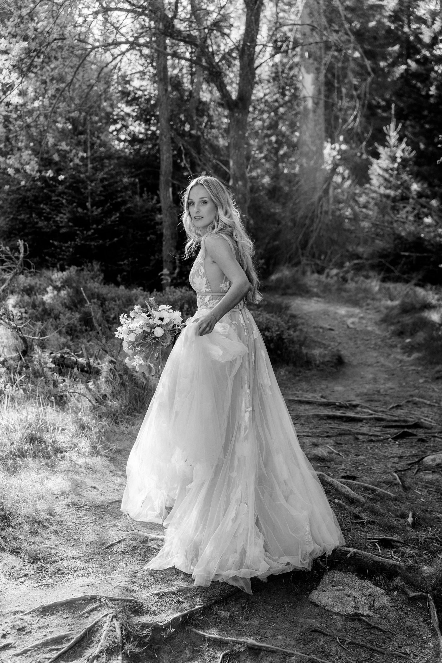 A bride in a flowing white lace gown stands gracefully on a forest path holding a bouquet in black and white photography.