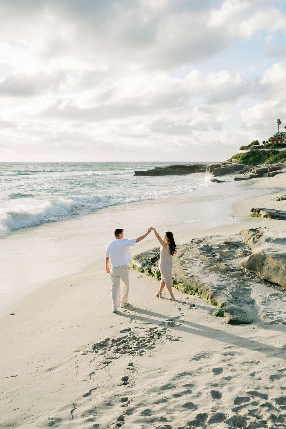 Couple walking hand in hand along a scenic beach with rocky cliffs and crashing waves during sunset.