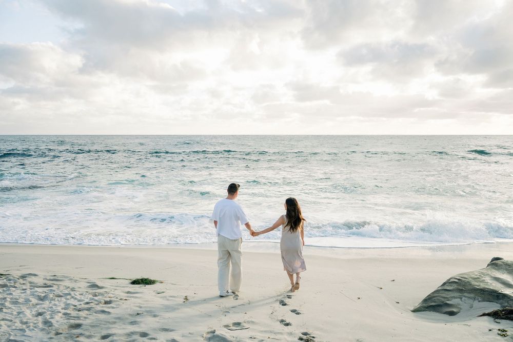 A couple strolls hand in hand along a serene beach with crashing waves and cloudy skies in the background.