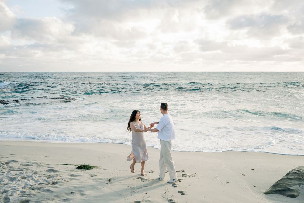 Two people in white outfits dancing together on a beautiful beach at sunset with turquoise waves crashing nearby.