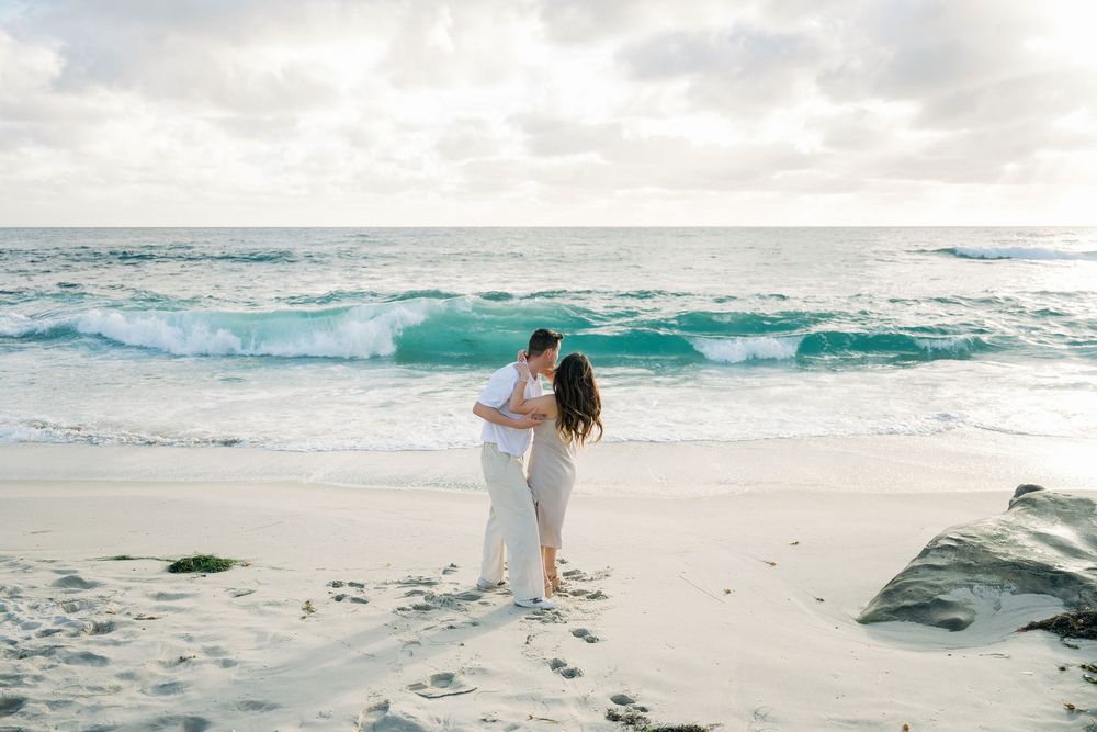 A couple embraces while standing on a pristine white sand beach with turquoise waves rolling in under cloudy skies.