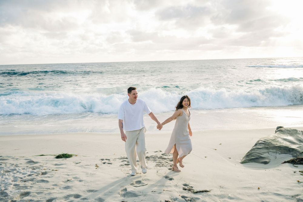 Two people in white and beige outfits walk together on a sandy beach beside the rolling ocean waves.