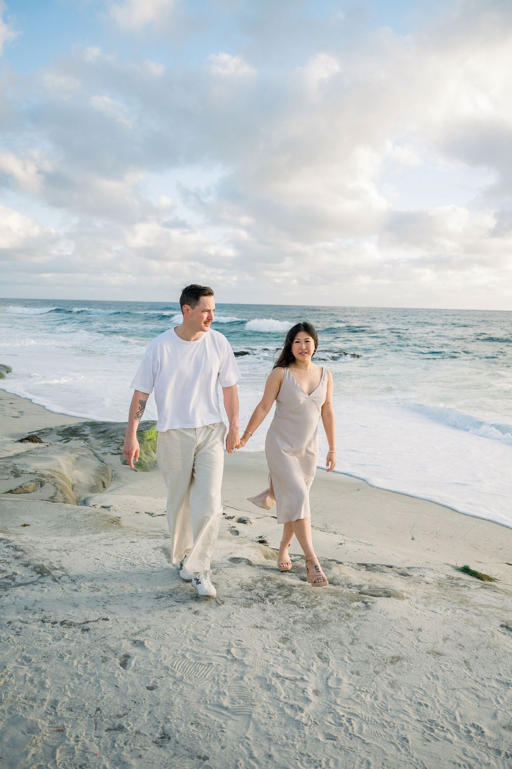 A couple walking hand in hand on a scenic beach at sunset with waves crashing against the shore in the background.